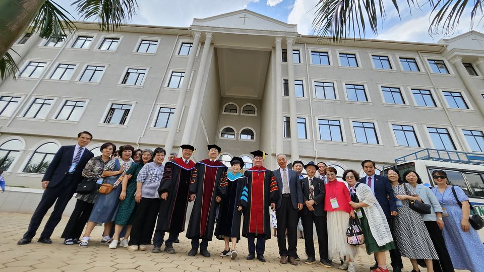 A large group, some in academic robes, standing in front of a white multi-storey building